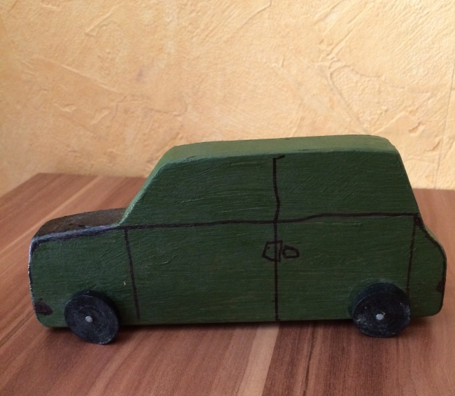 Aus Holz ausgesägtes und grün-schwarz lackiertes Auto mit schwarzen, beweglichen Reifen und aufgemalten Türen und Fenstern